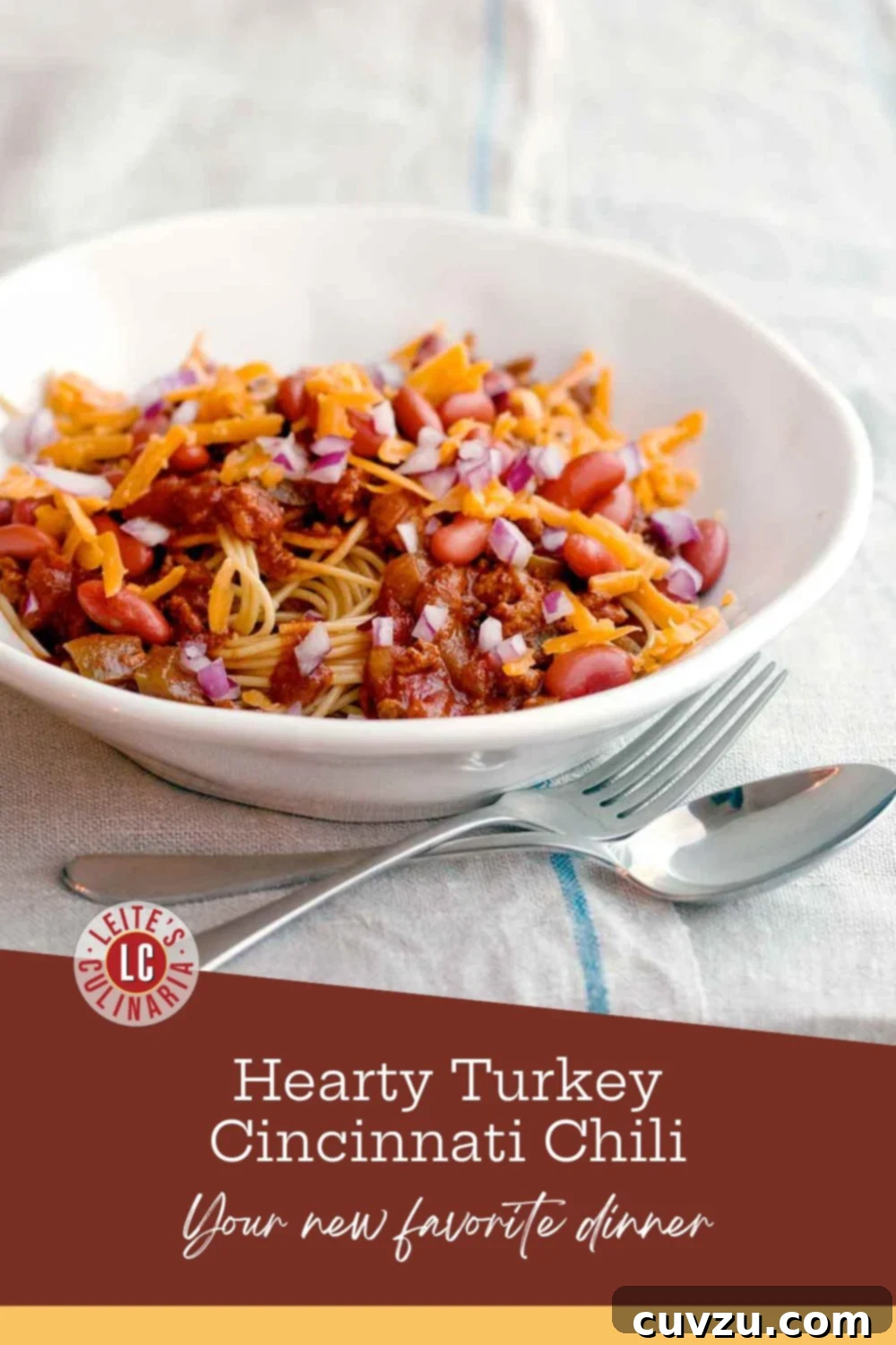 A white bowl of turkey Cincinnati chili with shredded cheese and diced red onion over spaghetti, with a fork and spoon on the table.