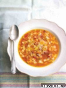 A white bowl filled with pasta soup with potatoes and pancetta on a linen cloth with a spoon resting on the edge of the bowl.