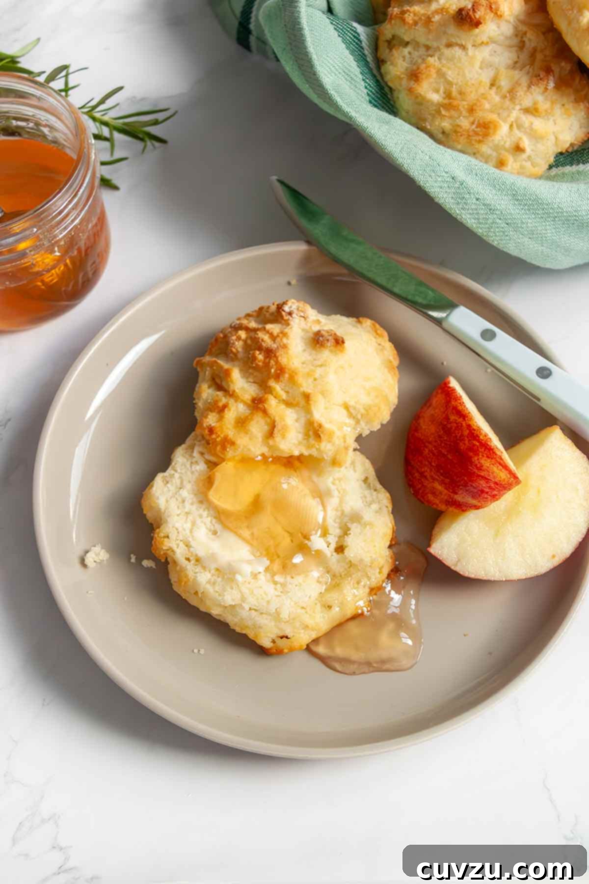 A freshly cut buttermilk drop biscuit on a plate, topped with shimmering apple jelly, with a knife and a few apple wedges on the side.