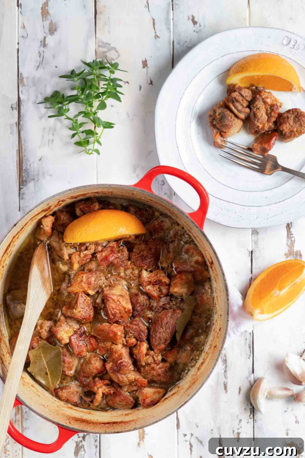 Carnitas: Authentic Mexican Braised Pork 7 A red Dutch oven filled with braised pork cubes with an orange wedge on top and a few orange wedges, garlic cloves, oregano, and spoon on the side.