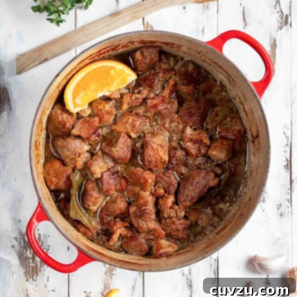 A red Dutch oven filled with braised pork cubes with an orange wedge on top and a few orange wedges, garlic cloves, oregano, and spoon on the side.