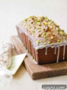 A pistachio pound cake on a wooden cutting board, garnished with a light glaze and crushed pistachios, with whole pistachios scattered nearby.