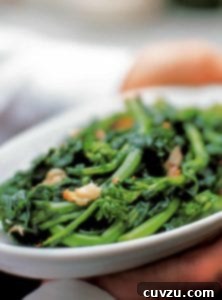 A white platter filled with vibrant green broccoli rabe, generously topped with golden roasted garlic cloves and a sprinkle of red pepper flakes.