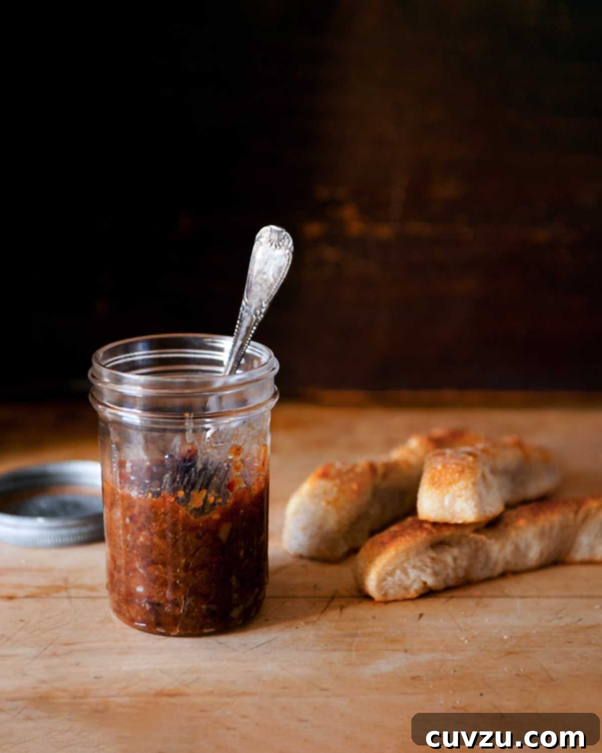Smoky Sweet Bacon Reserve 4 A charming jar of homemade bacon jam with a spoon resting inside, placed on a rustic wooden board, with a lid and breadsticks softly blurred in the background.
