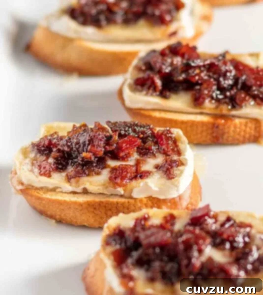 Four crostini topped with Brie and bacon jam, arranged on a rustic board for an appetizer.
