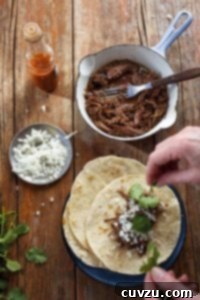 A person assembling a slow cooker barbacoa taco with a pot of shredded barbacoa meat, a small dish of onions, a bottle of hot sauce, and some cilantro around the taco plate.