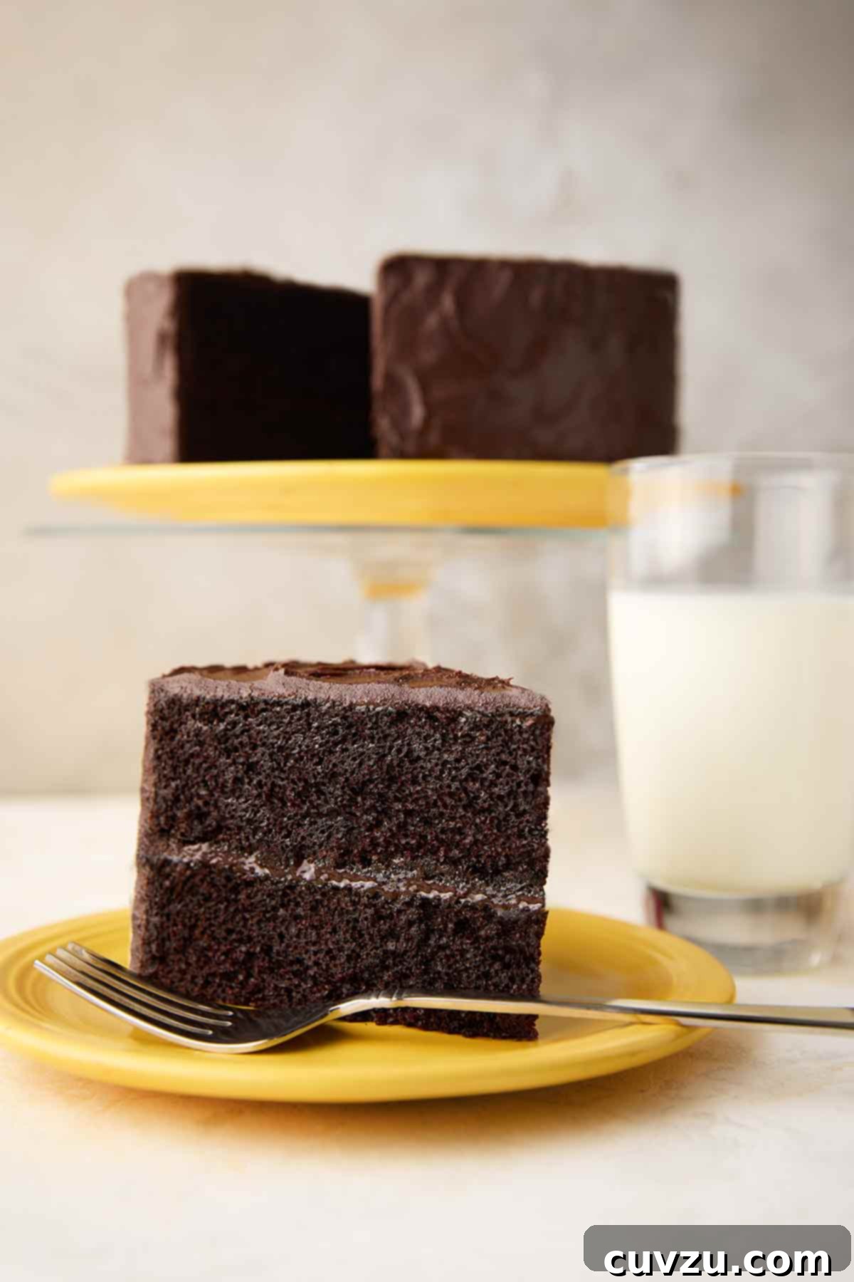 Next-Level Hershey's Chocolate Cake 9 A perfectly portioned slice of Hershey's chocolate cake, generously covered in creamy chocolate frosting, rests on a vibrant yellow plate. In the background, the remaining elegant cake sits invitingly on a cake stand, promising more delicious moments.