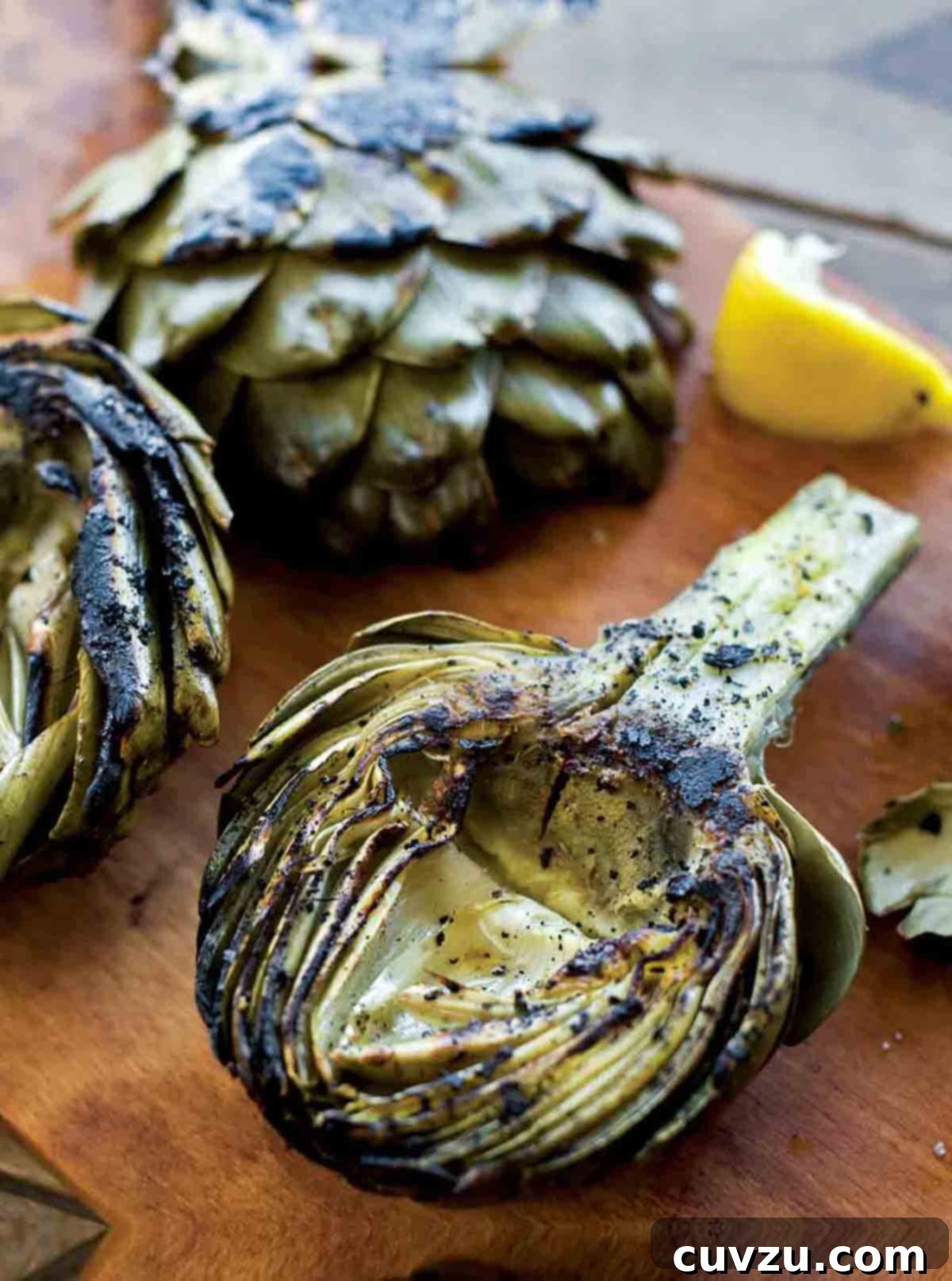 Flame-Kissed Artichoke Hearts 2 Three grilled artichoke halves, some lemon wedges, and a creamy dip served on a rustic wooden board, ready for a delightful summer meal.