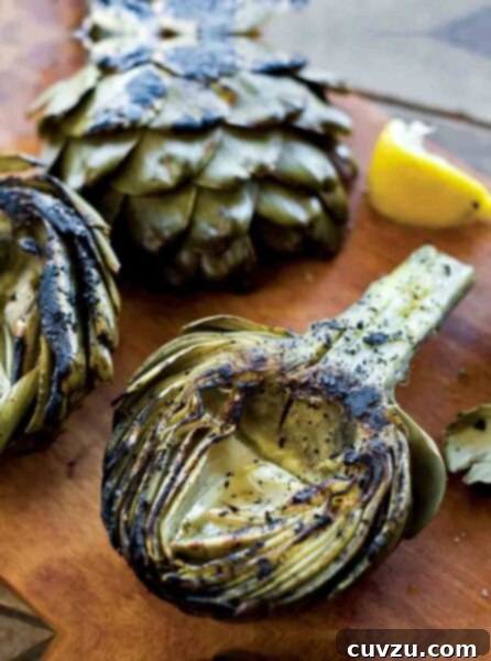 Three grilled artichoke halves, some lemon wedges, and a creamy dip on a wooden peel, highlighting the perfect char and tender texture.