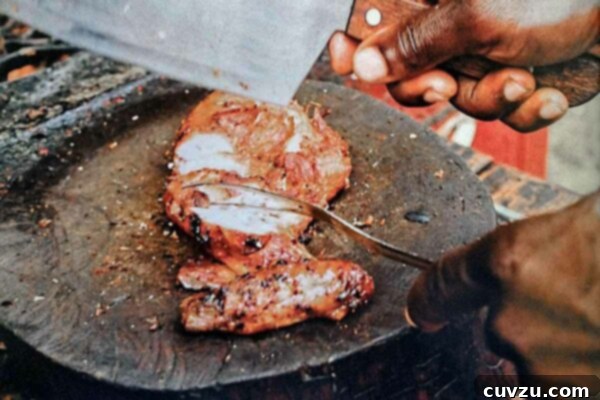 A sliced jerk chicken breast on a round cutting board, with a man's hand holding a cleaver above it