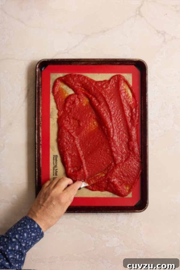 The Art of Homemade Tomato Paste 9 A hand using a small spatula to spread thick tomato purée onto a silicone mat on a baking sheet.