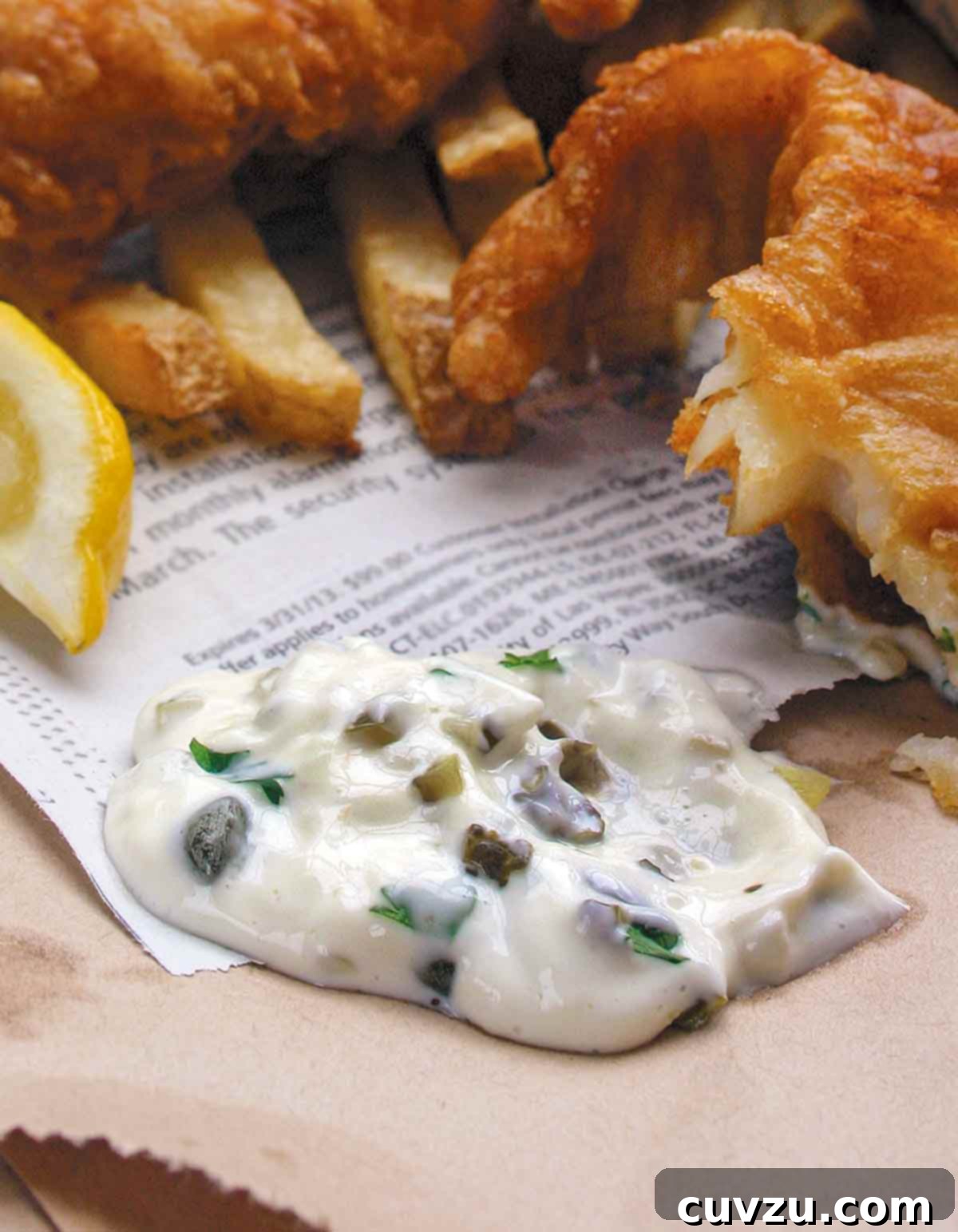 A dollop of homemade tartar sauce on a piece of newspaper with fried fish and a lemon wedge beside it.