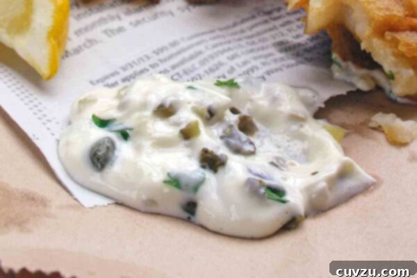 A dollop of homemade tartar sauce on a piece of newspaper with fried fish and a lemon wedge beside it.