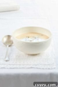 A white bowl of creamy cauliflower and blue cheese soup on a white linen cloth with a spoon resting beside it, garnished with a sprinkle of blue cheese.