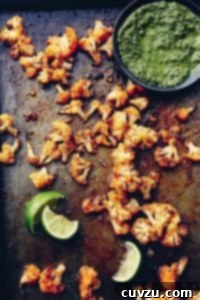 Florets of spicy roasted cauliflower on a baking sheet with squeezed lime wedges and a bowl of green sauce beside them.