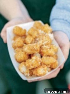 Two hands holding a paper container of tater tots generously sprinkled with grated Parmesan cheese.