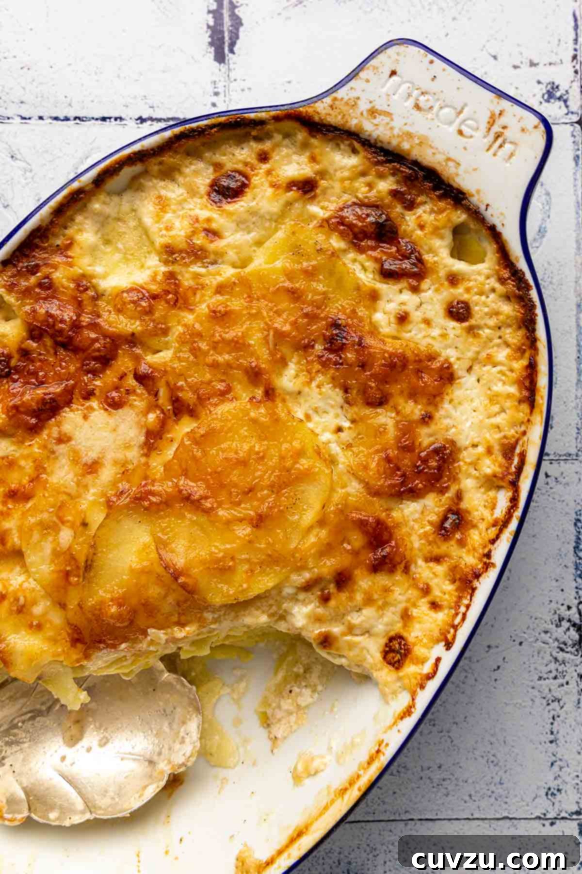 An oval casserole dish filled with golden-brown potatoes dauphinoise, with a silver serving spoon resting gently in the creamy layers, ready to be enjoyed.