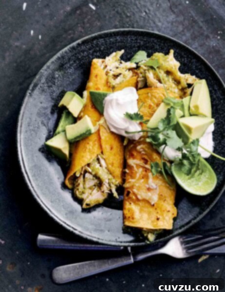 Two chicken enchiladas with green sauce in a black shallow bowl with cubed avocado, sour cream, cilantro, and a lime half.