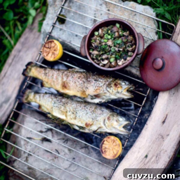 Two whole grilled trout on a grate over an open fire with a dish of mushrooms and two grilled lemon halves on the side.