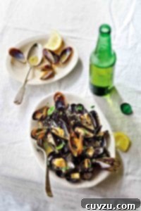 White plate with steamed mussels in beer, topped with parsley, nearby lemon wedges and a bottle of beer
