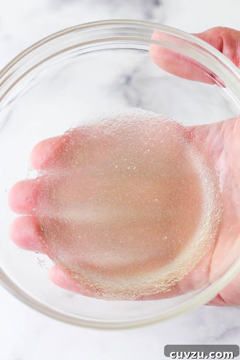 Bloom gelatin by combining with remaining cold champagne. A hand gently sprinkling powdered gelatin over cool champagne in a small bowl.