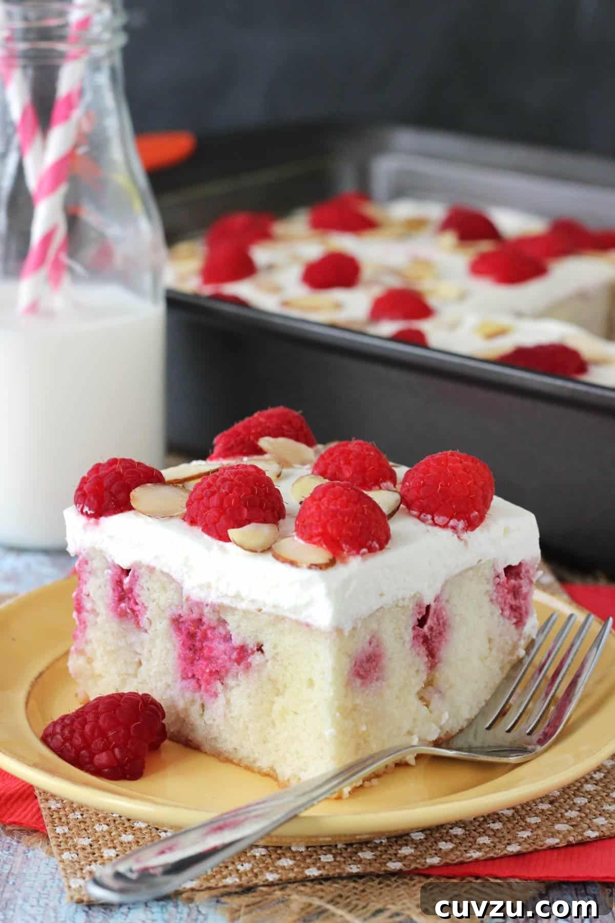 Raspberry Almond Infusion Cake 4 A beautiful close-up of a slice of Raspberry Almond Poke Cake on a yellow plate, with the full cake visible in the blurred background, highlighting its rich texture and topping.