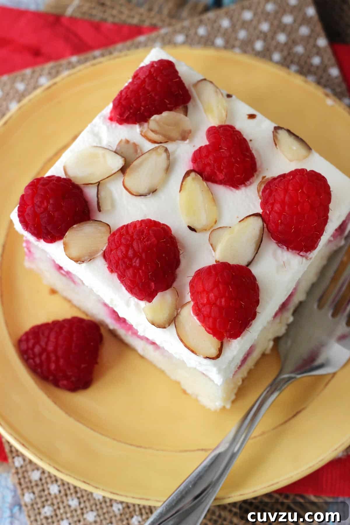 Raspberry Almond Infusion Cake 5 Top-down view of a generous slice of Raspberry Almond Poke Cake on a yellow plate, showing the creamy whipped topping and vibrant raspberry swirls.