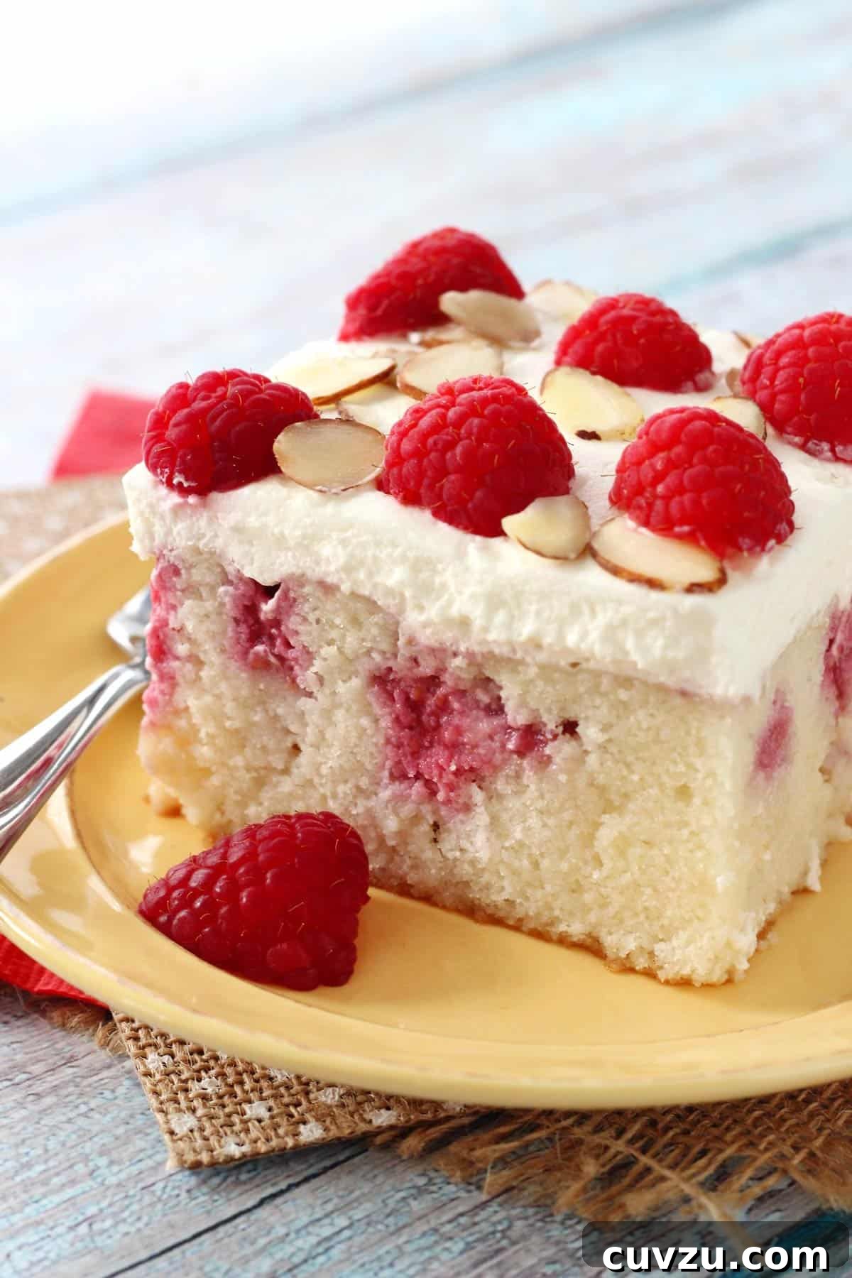 Raspberry Almond Infusion Cake 6 A detailed close-up of a slice of Raspberry Almond Poke Cake on a yellow plate, revealing the moist cake, berry filling, and luscious whipped cream.
