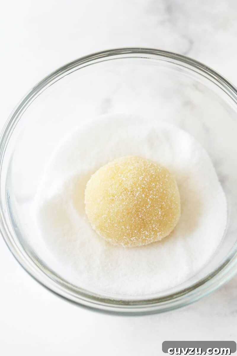 Achieve the perfect texture and appearance by rolling delicate balls of lemon cookie dough first in granulated sugar. The process of rolling a ball of vibrant lemon cookie dough in a bowl of granulated sugar, preparing it for its iconic crackle.
