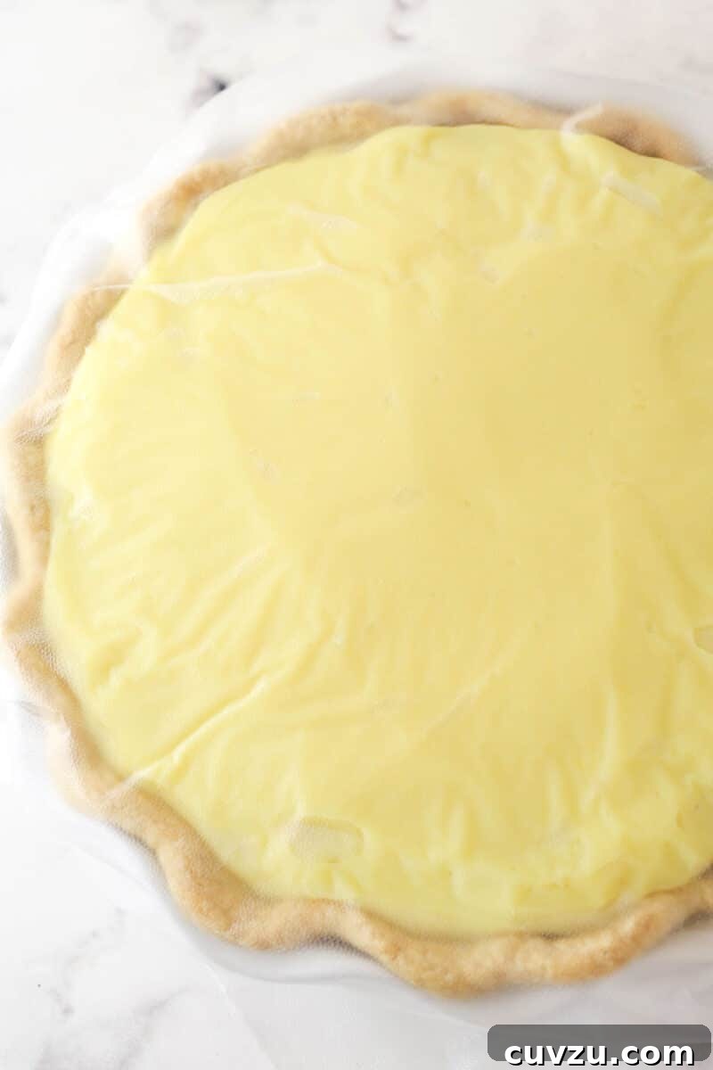 The prepared banana pudding pie, chilling with plastic wrap pressed directly onto the surface. The banana pudding pie, fully assembled with its layers, covered directly with plastic wrap for chilling.
