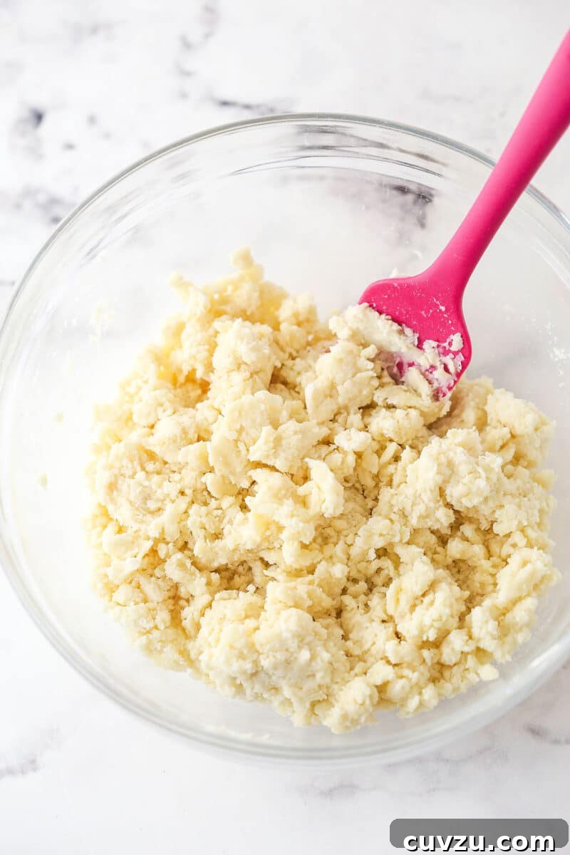 Combining pie crust ingredients: adding ice water and folding until the dough forms. The pie crust dough being gently folded with a spatula in a mixing bowl, showing the gradual incorporation of ice water.