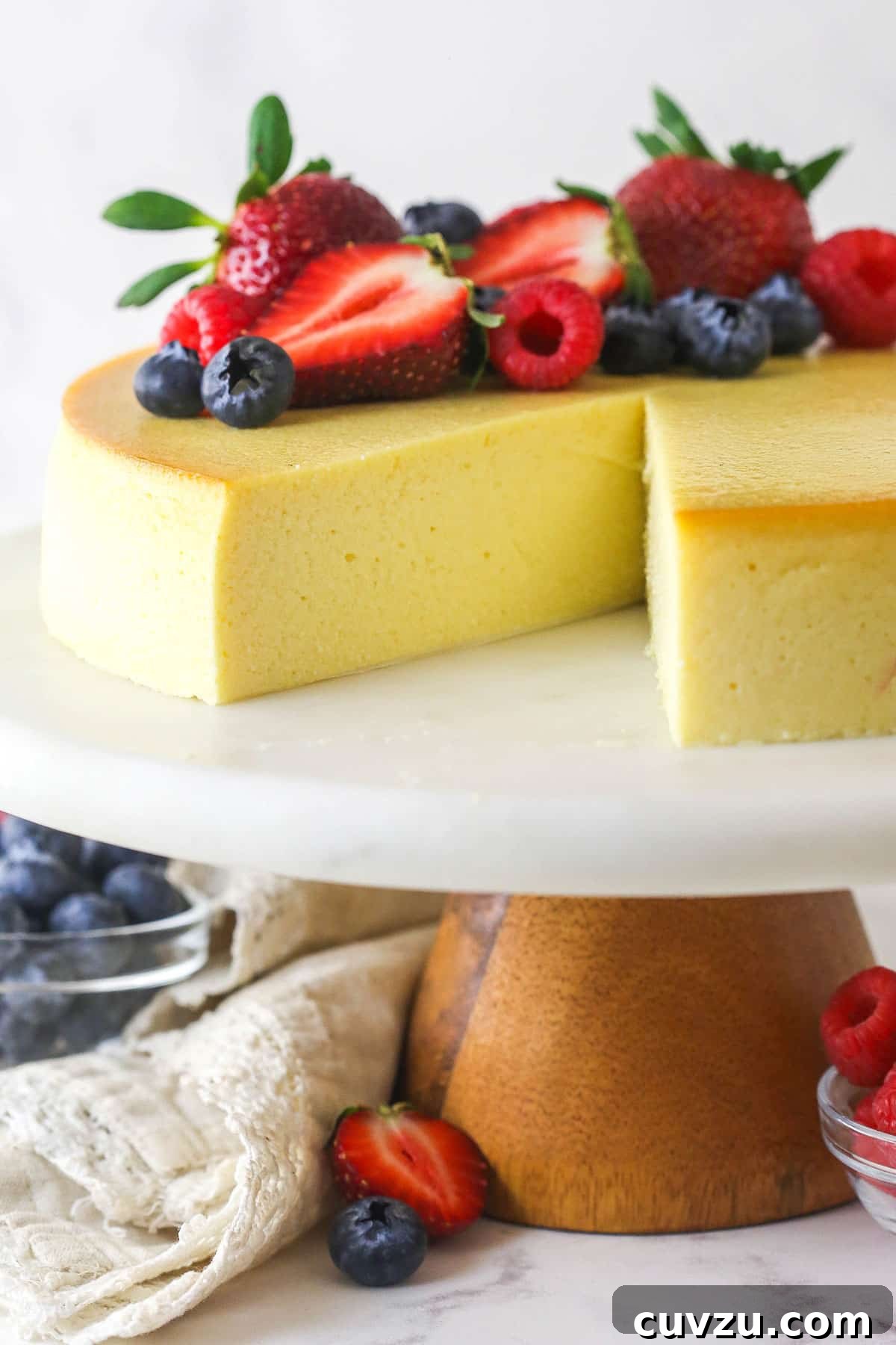 Easy 5-Ingredient Ricotta Cheesecake with Lemon Zest A whole ricotta cheesecake on a elegant cake stand with one slice removed, revealing its creamy texture. Topped with a sprinkle of powdered sugar and fresh berries.