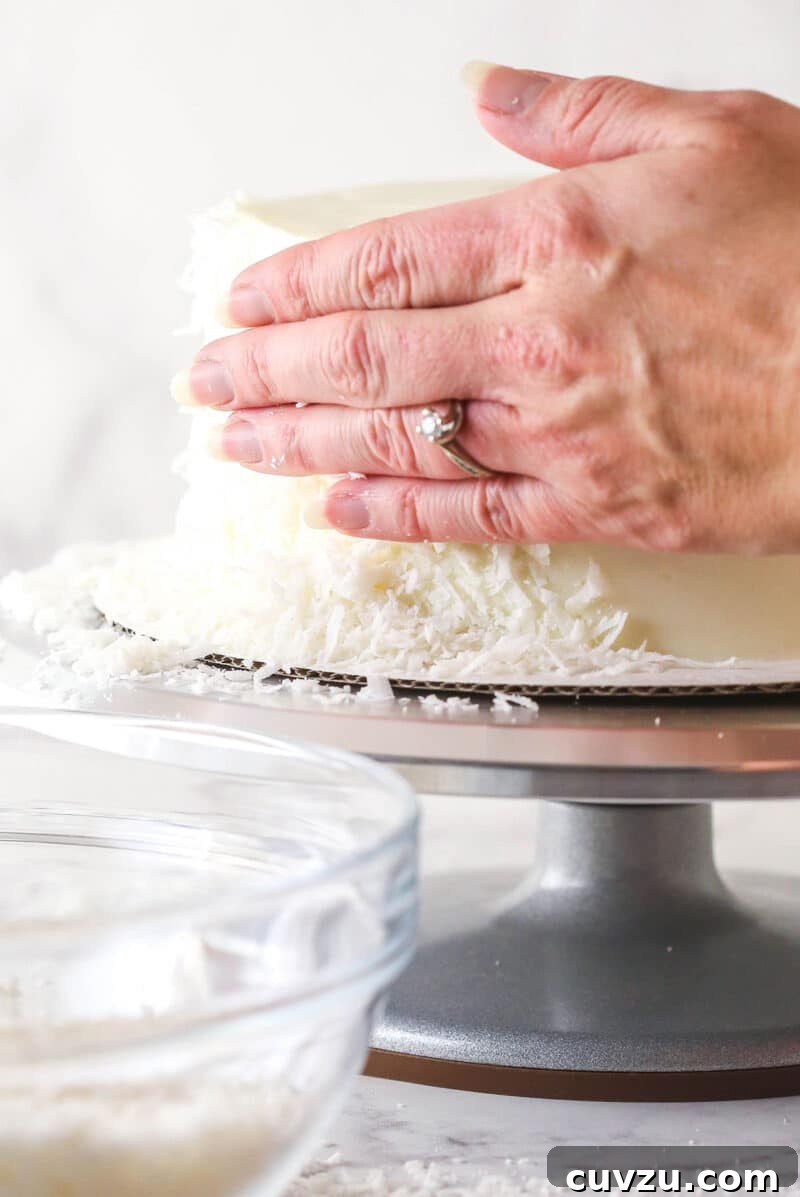 Tropical Delight Cake 14 Shredded coconut being pressed gently onto the side of a frosted cake on a turntable.