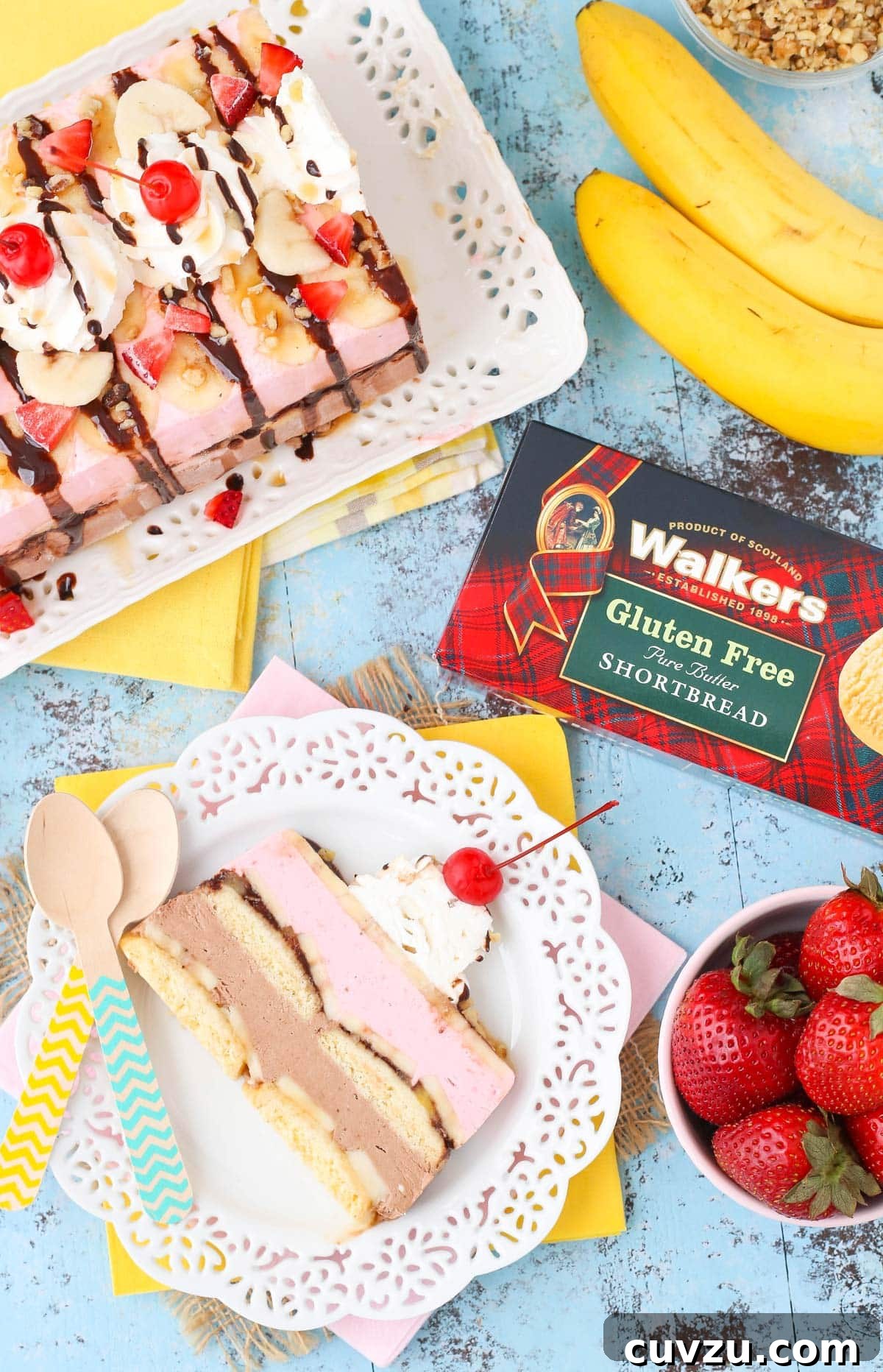 Frozen Banana Split Loaf 4 An overhead shot of a single slice of banana split ice cream cake loaf on a white plate, garnished with fresh fruit, nuts, and a cherry, accompanied by whole cookies and additional toppings.