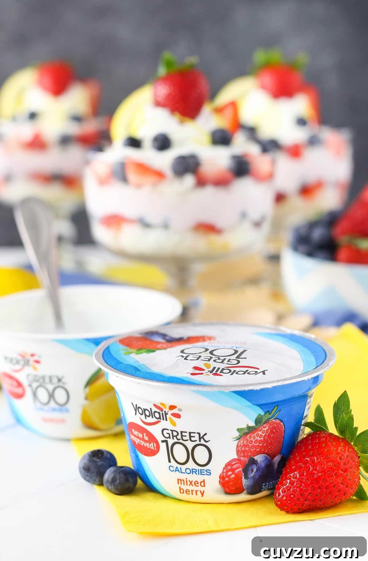 Tangy Berry Bliss Layers 6 A container of Yoplait Greek 100 yogurt in the foreground, with blurred lemon berry yogurt trifles in the background, emphasizing the yogurt's role.