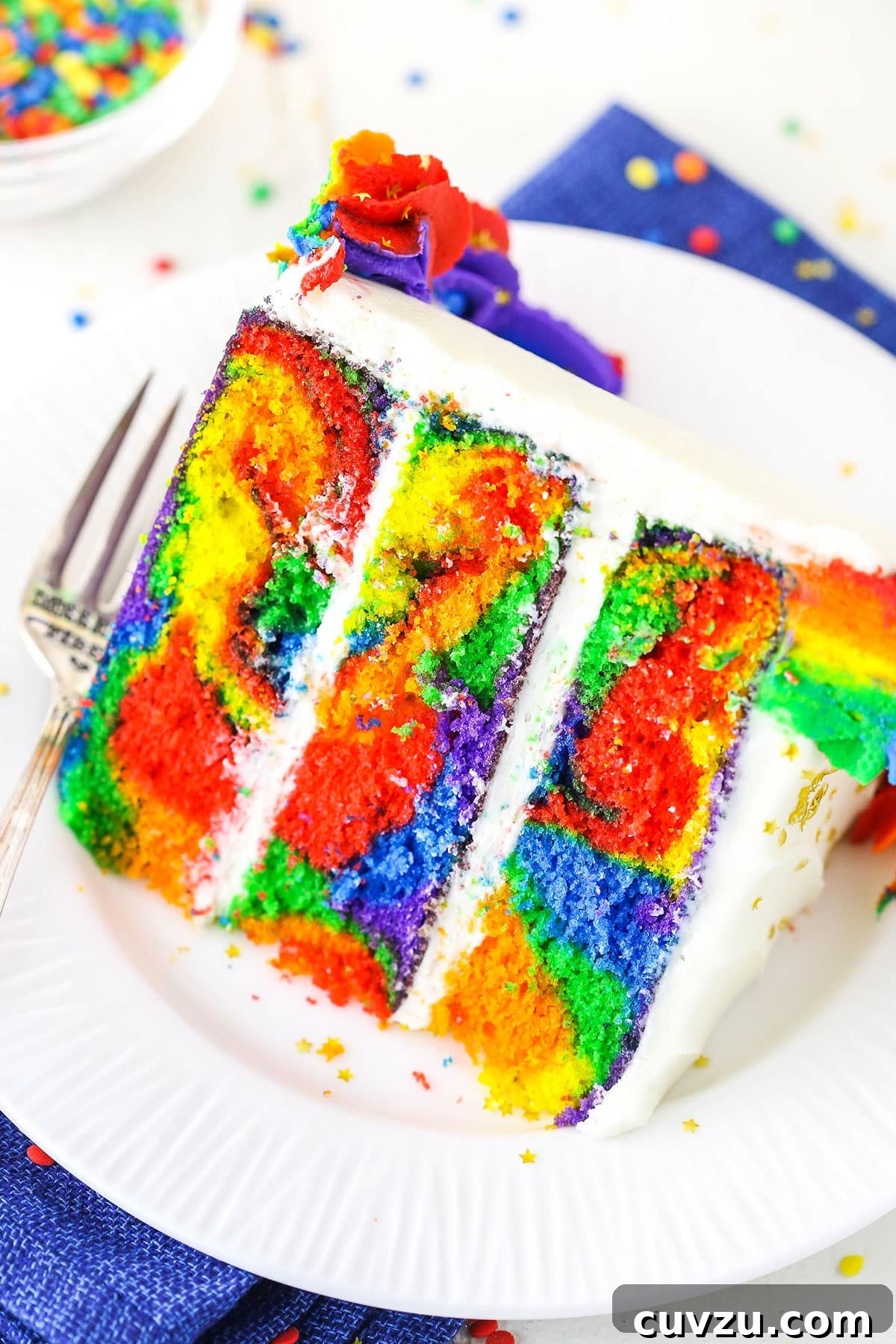 Kaleidoscope Cake 2 A perfectly sliced Rainbow Swirl Cake revealing vibrant internal layers next to a silver fork on a clean white plate, ready to be enjoyed.