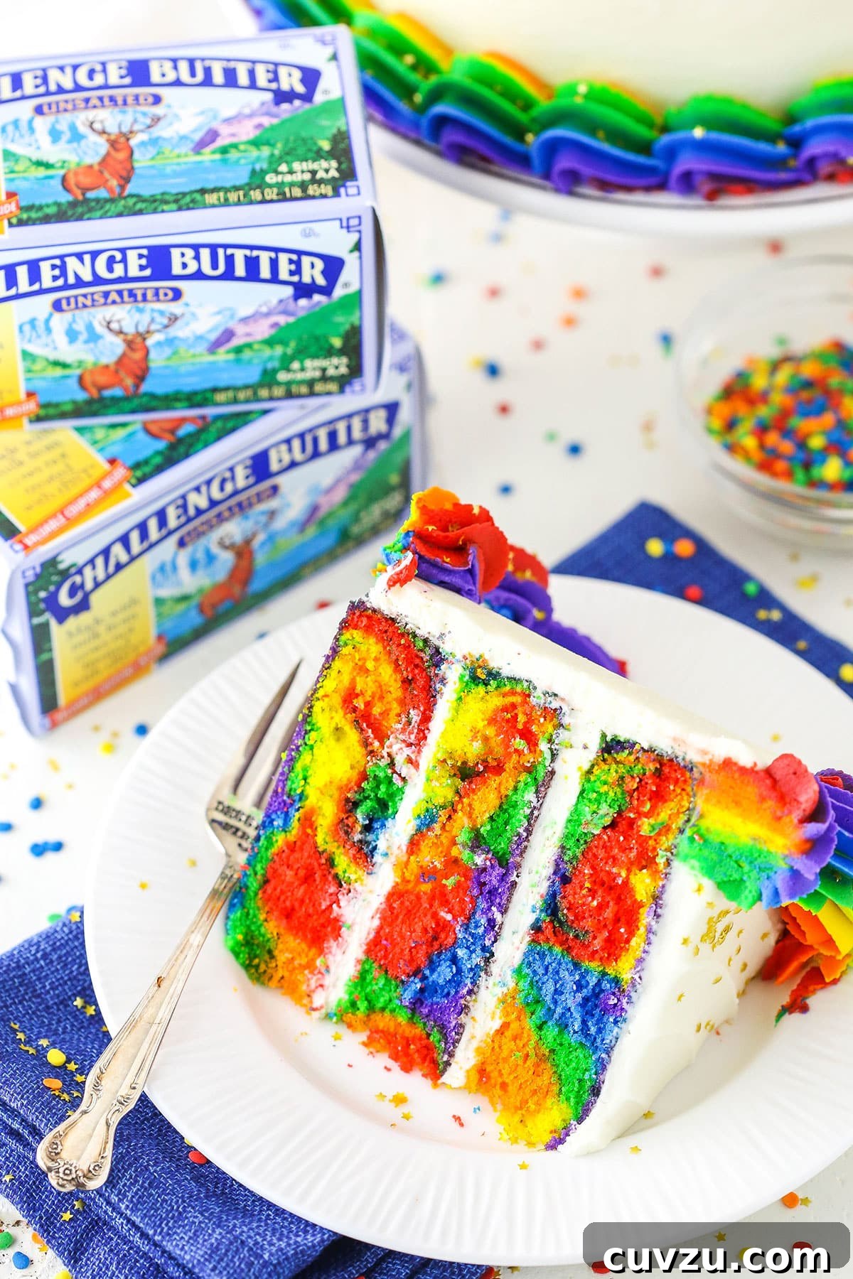 Kaleidoscope Cake 5 A vibrant slice of Rainbow Swirl Cake rests beside a fork on a white plate, with Challenge Butter boxes softly blurred in the background, hinting at the quality ingredients.