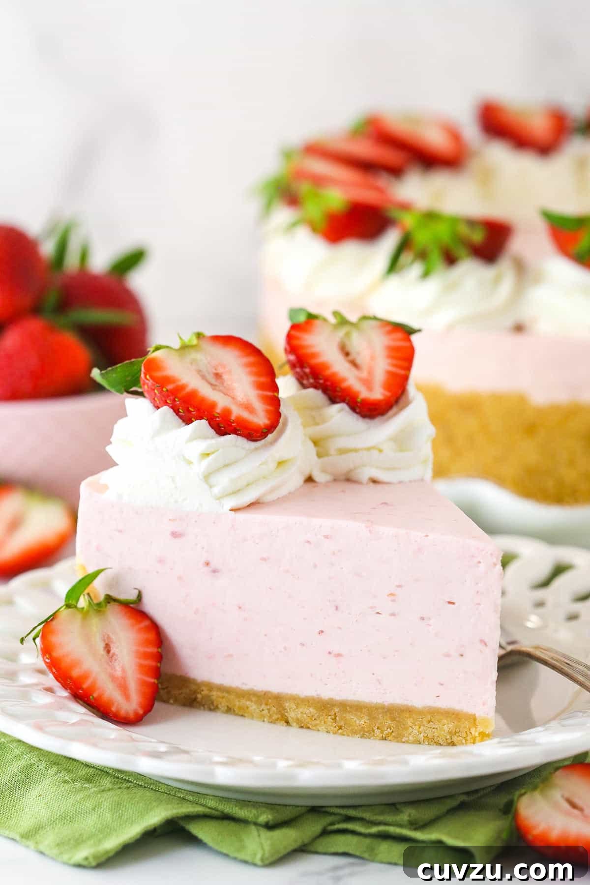 This no-bake strawberry cheesecake features a rich vanilla wafer crust, a smooth strawberry cream cheese filling, and a homemade whipped cream topping, garnished with fresh strawberries. A beautifully presented slice of no-bake strawberry cheesecake on a white plate, with a fork poised to take a bite. In the background, a whole no-bake strawberry cheesecake is visible next to a bowl of fresh, ripe strawberries, highlighting the dessert's fresh ingredients and appeal.