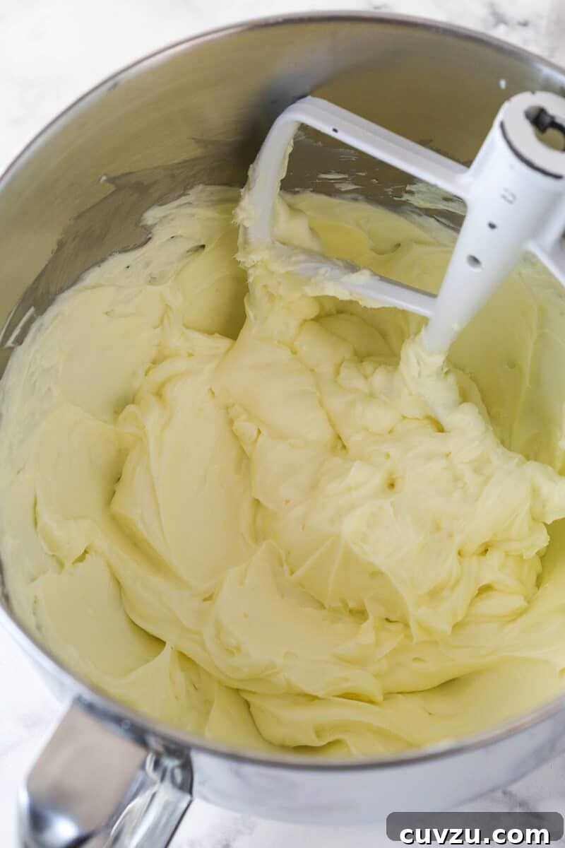 Beating room temperature cream cheese and sugar until smooth for the no-bake strawberry cheesecake filling. Cream cheese and sugar being beaten together in a large mixer bowl with an electric hand mixer until the mixture is light, fluffy, and perfectly smooth, demonstrating a key step in creating the cheesecake base.