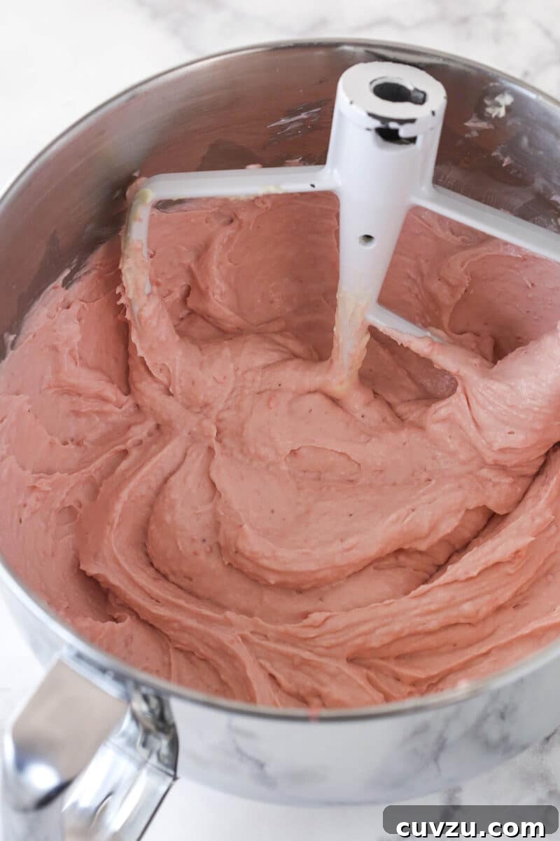 Gradually beating the strawberry mixture into the cream cheese and sugar for a smooth no-bake cheesecake filling. The vibrant red strawberry mixture being gradually beaten into the creamy white cream cheese and sugar mixture in a large bowl, ensuring a uniform and smooth cheesecake filling.