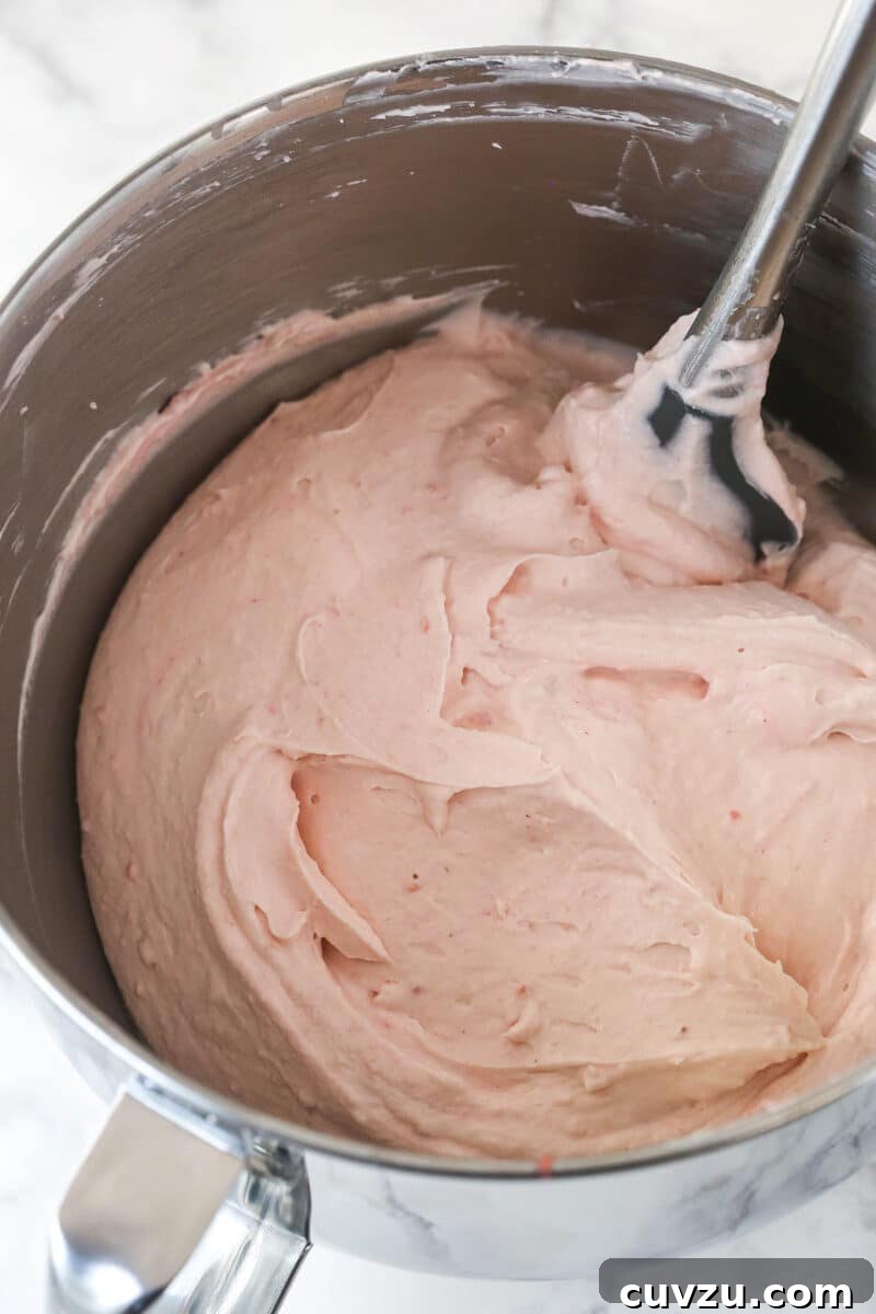 Gently folding whipped cream into the strawberry cream cheese mixture for the no-bake cheesecake filling. A rubber spatula gently folding the freshly whipped cream into the strawberry cream cheese mixture, ensuring a light and airy texture for the no-bake cheesecake filling.