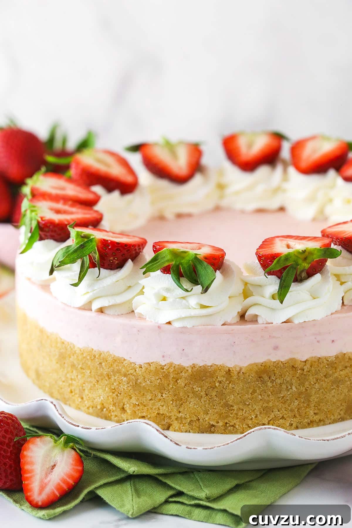 This no-bake strawberry cheesecake is made of a buttery vanilla wafer crust, a creamy strawberry filling, and homemade whipped cream, topped with fresh berries. A complete no-bake strawberry cheesecake, beautifully adorned with whipped cream swirls and fresh sliced strawberries, presented on a serving platter next to more fresh strawberries, ready to be enjoyed.