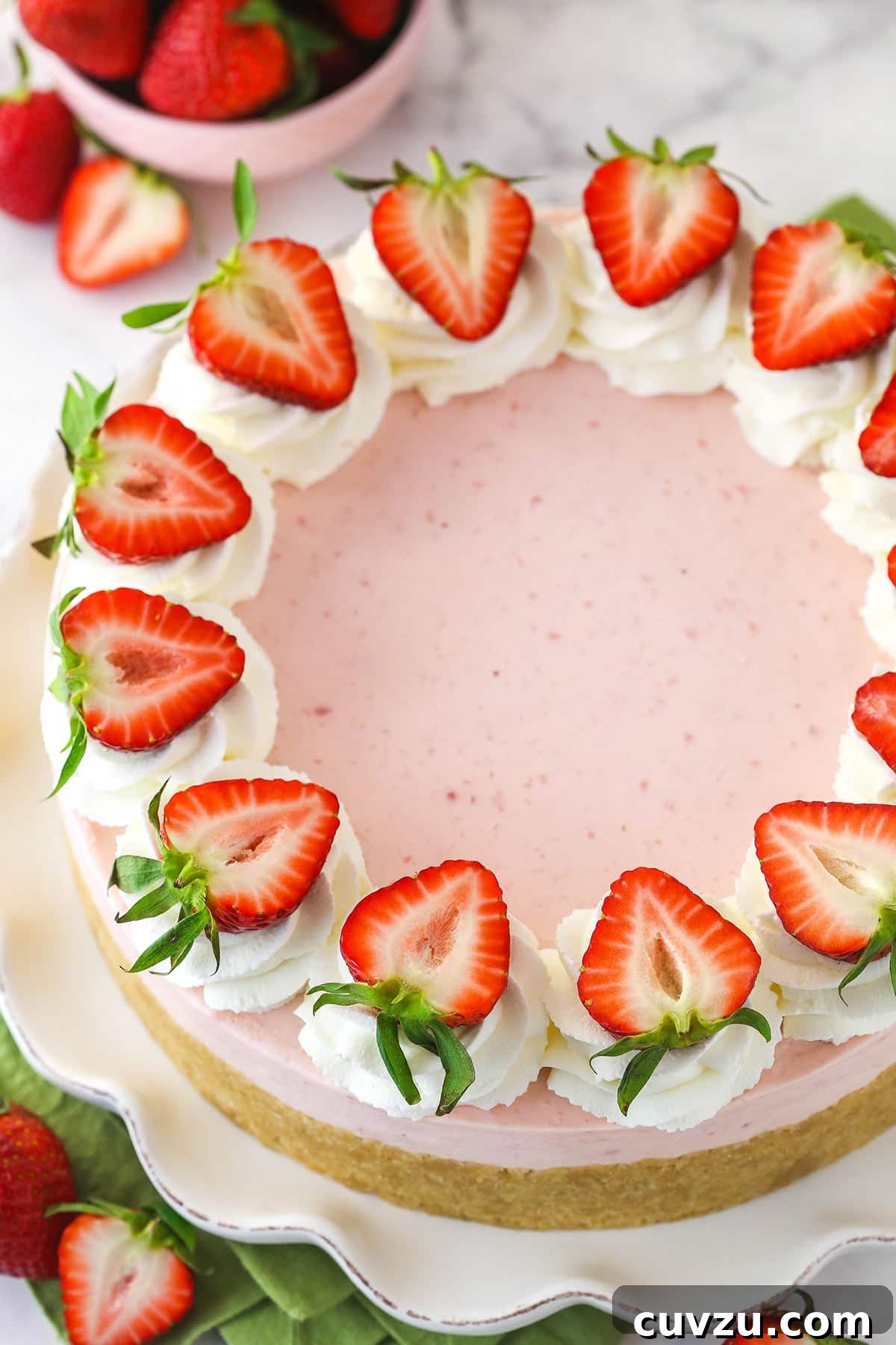 This no-bake strawberry cheesecake features a buttery vanilla wafer crust, a creamy strawberry filling, and a generous topping of homemade whipped cream, all made without baking. An overhead view of a complete no-bake strawberry cheesecake, elegantly presented on a serving platter and surrounded by an assortment of fresh, whole strawberries. The vibrant red of the berries contrasts beautifully with the creamy white topping, making it an inviting dessert.