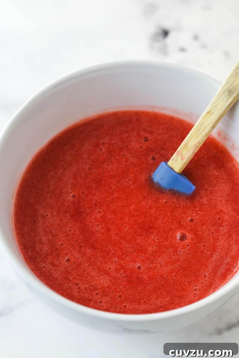 Freshly made strawberry puree, ready to be cooked for the no-bake strawberry cheesecake filling. Smooth, vibrant strawberry puree in a glass bowl, with a rubber spatula resting inside, indicating the successful processing of fresh strawberries for the cheesecake filling.