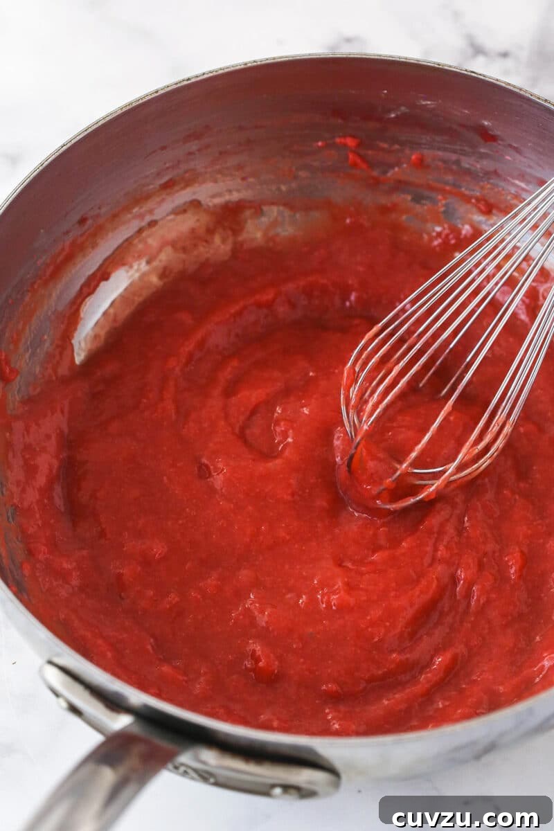 Whisking the strawberry mixture with cornstarch to thicken it for the no-bake cheesecake filling. A hand stirring a thickening strawberry mixture in a saucepan with a whisk over medium heat, showing the cooking process for the strawberry component of the cheesecake filling.