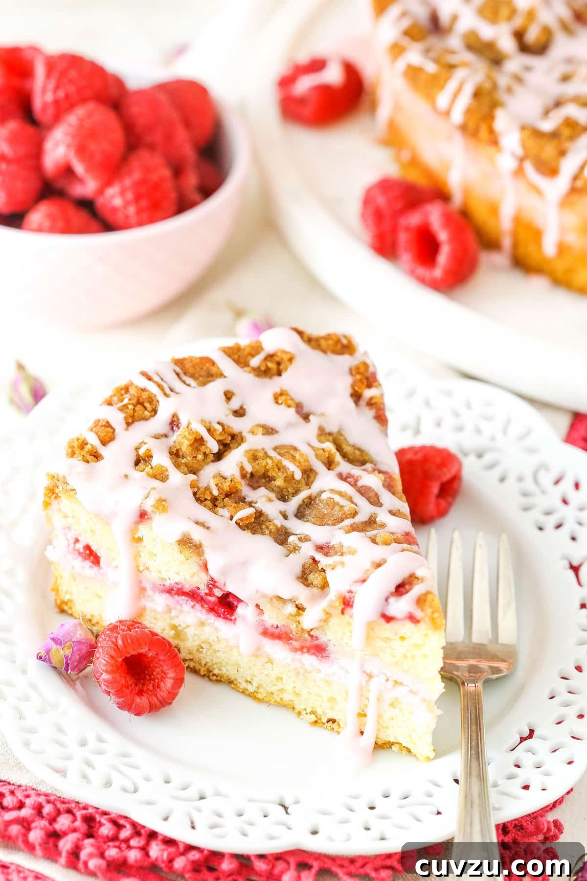 Each slice of this Raspberry Rose Coffee Cake offers a delightful combination of vanilla rose cream cheese, fresh raspberries, and a crumbly streusel. A perfect slice of Raspberry Rose Coffee Cake on a white plate, revealing its layers of moist cake, creamy filling, and fresh raspberries, with a fork resting beside it.