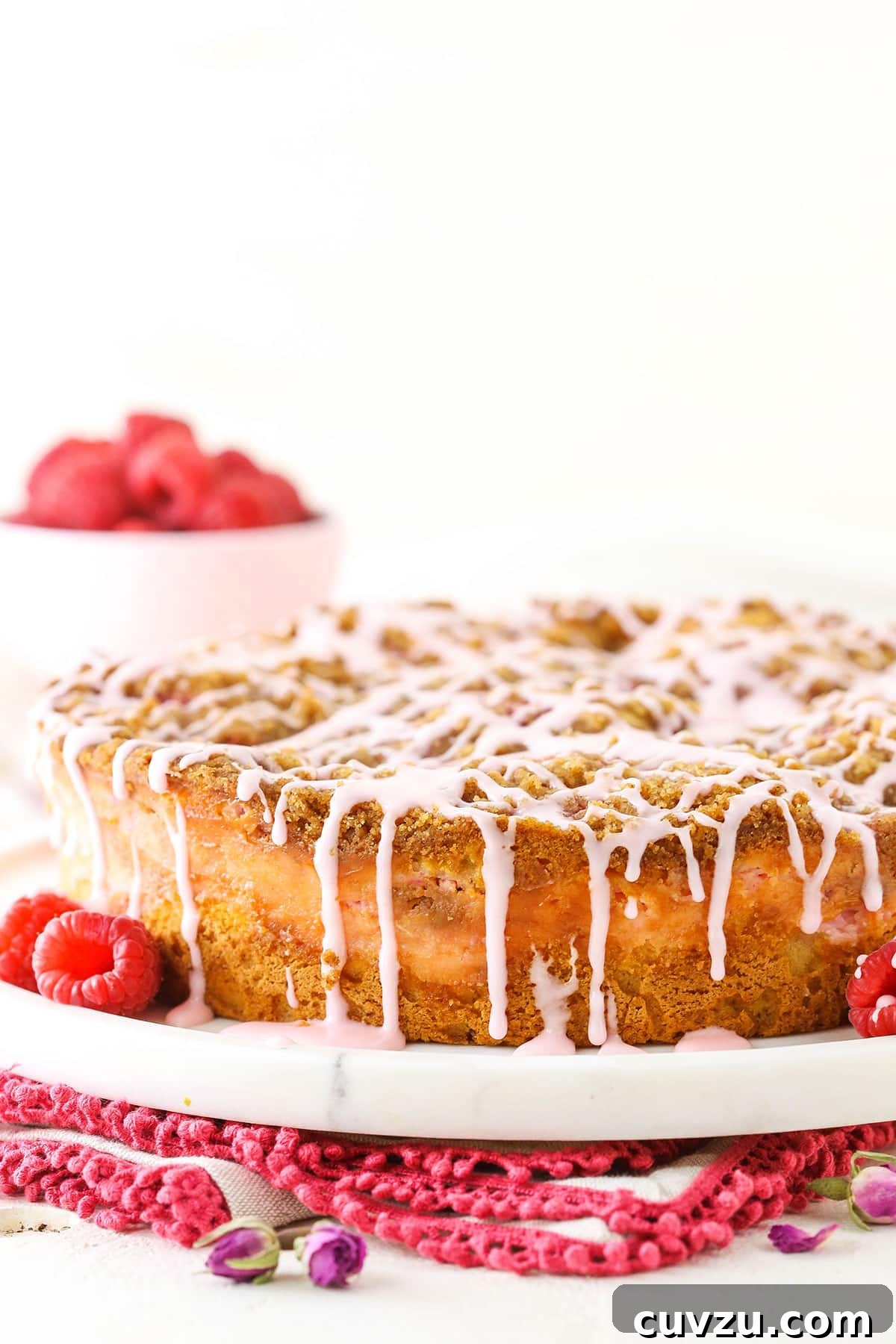 The multi-layered Raspberry Rose Coffee Cake features a delicious streusel and a beautiful rose glaze. A stunning side view of the Raspberry Rose Coffee Cake, showcasing its golden-brown streusel topping, pink glaze, and the vibrant red raspberries peaking through the layers, on a pristine white serving platter.