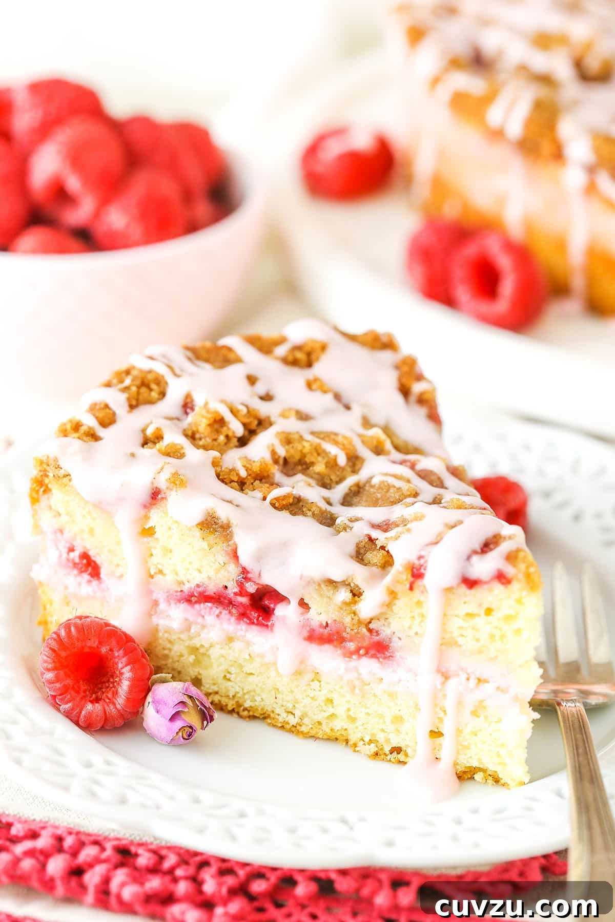 Enjoy a slice of Raspberry Rose Coffee Cake, featuring a delightful blend of creamy filling, juicy raspberries, and a crumbly topping. A delectable slice of Raspberry Rose Coffee Cake on a white plate with a fork, surrounded by fresh raspberries, highlighting its irresistible texture and vibrant fruit.