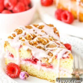 Velvet Rose Raspberry Coffee Cake 2 A slice of Raspberry Rose Coffee Cake next to a fork on a white plate with raspberries in the background