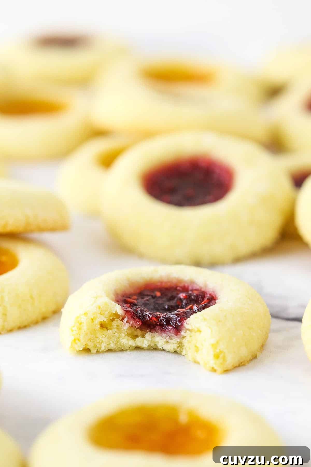 These classic thumbprint cookies are soft and chewy, filled with your favorite jam or preserves. Perfect for Christmas and easy to make! Close up of a jam thumbprint cookie with a bite missing, surrounded by more thumbprint cookies.